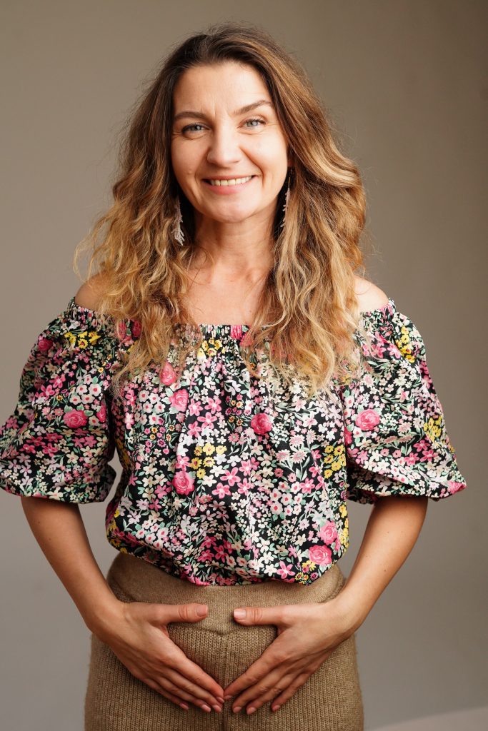 Adela Haru zâmbind, în bluză cu flori, cu mâinile pe pântec